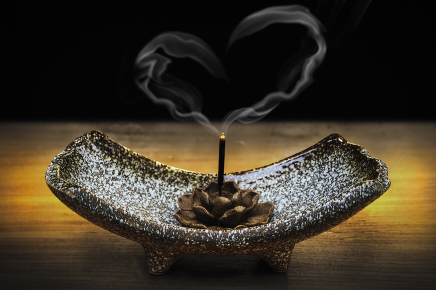 A burning incense stick in a metal flower base, letting off heart-shaped smoke