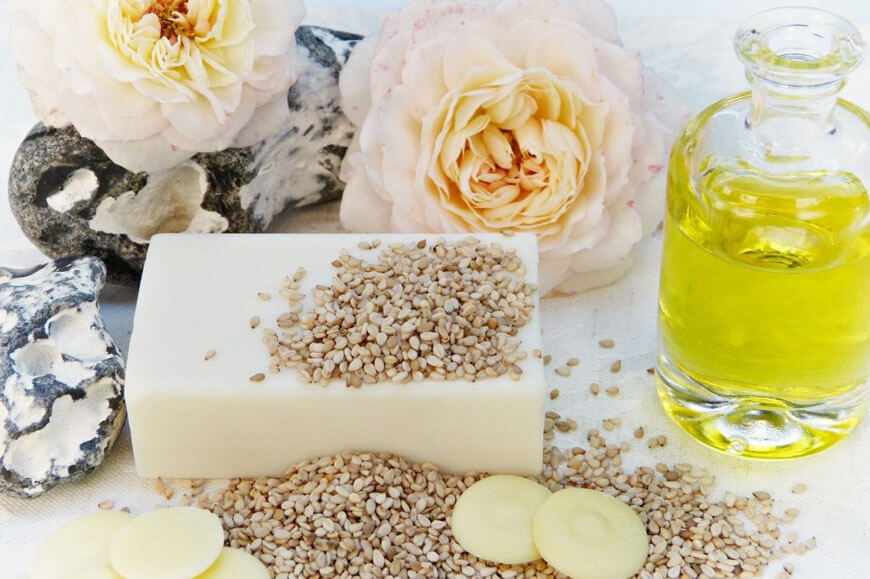 A white bar of soap with sesame seeds, roses, and a jar of oil beside it