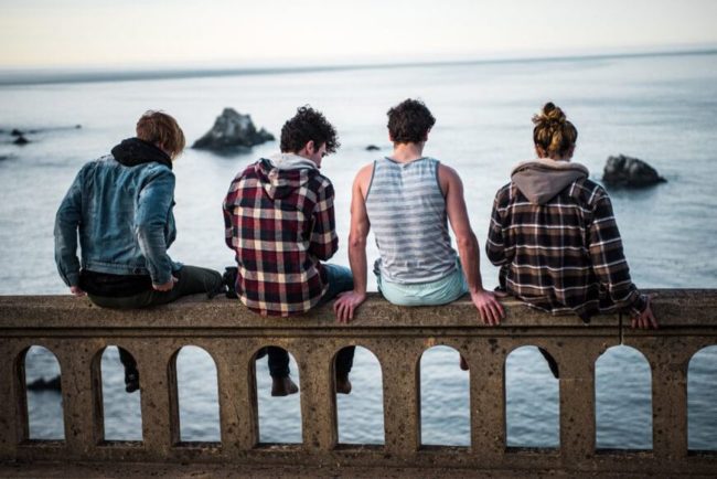 Four young people sitting on the edge of a bridge, looking out over the water