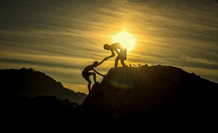 A man helping his friend up a mountain with sunset in the background