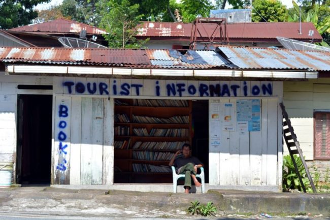 A man sitting on a chair outside a spray-painted shop front that reads “Tourist Information.”