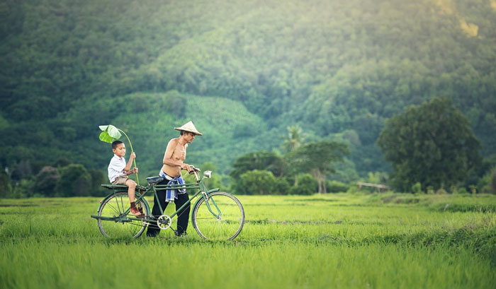 A Cambodian man pushing a bicycle through the grass with a boy seated on the back.