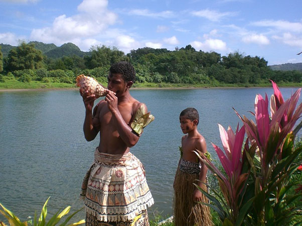 A native Fijian man and boy next to a river with a conch shell.