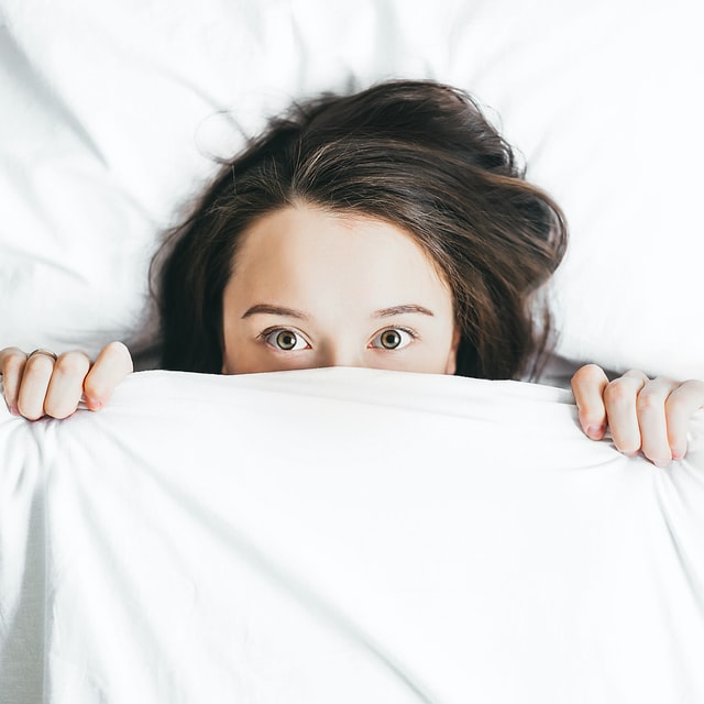 Woman with wide open eyes covers face with white comforter.