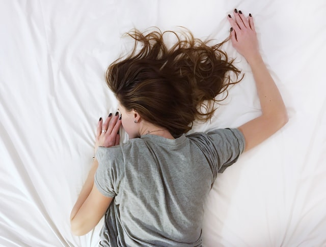Woman in grey t-shirt sleeps face down on white bed.